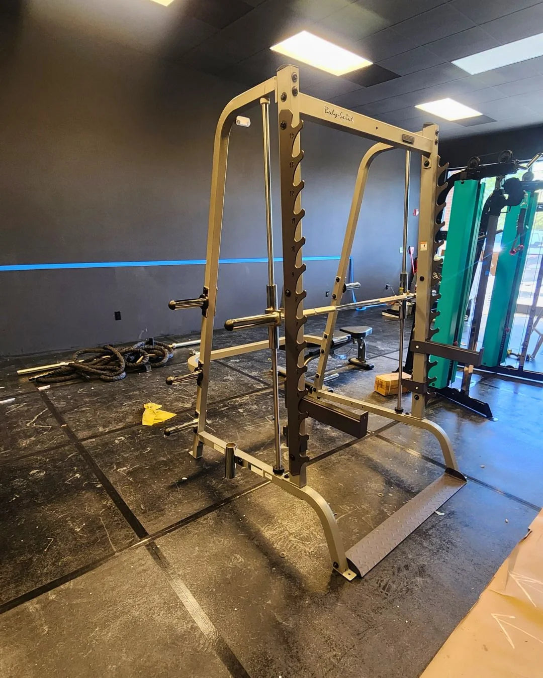 A gym with a Smith machine centered in the room. Theres a squat rack nearby, exercise ropes on the ground, and a green gym machine in the background. The walls are dark with a blue horizontal stripe. The floor is rubber matting.