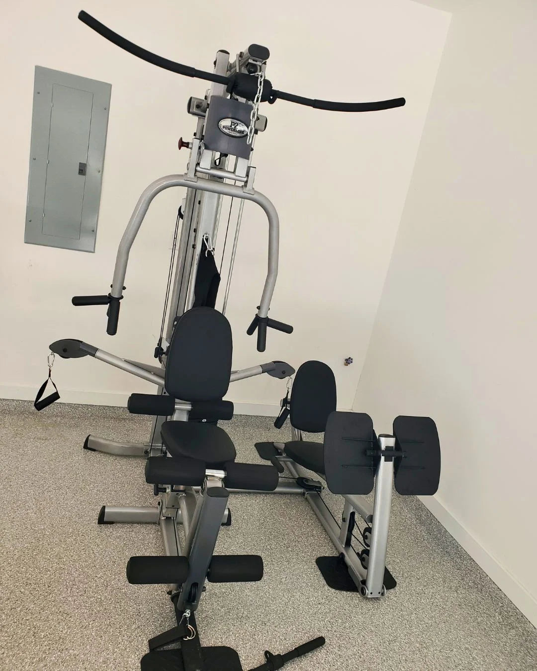 A multi-gym exercise machine with various attachments and seating is set up in a room with light-colored walls and a gray-speckled floor. An electrical panel is mounted on the wall nearby.