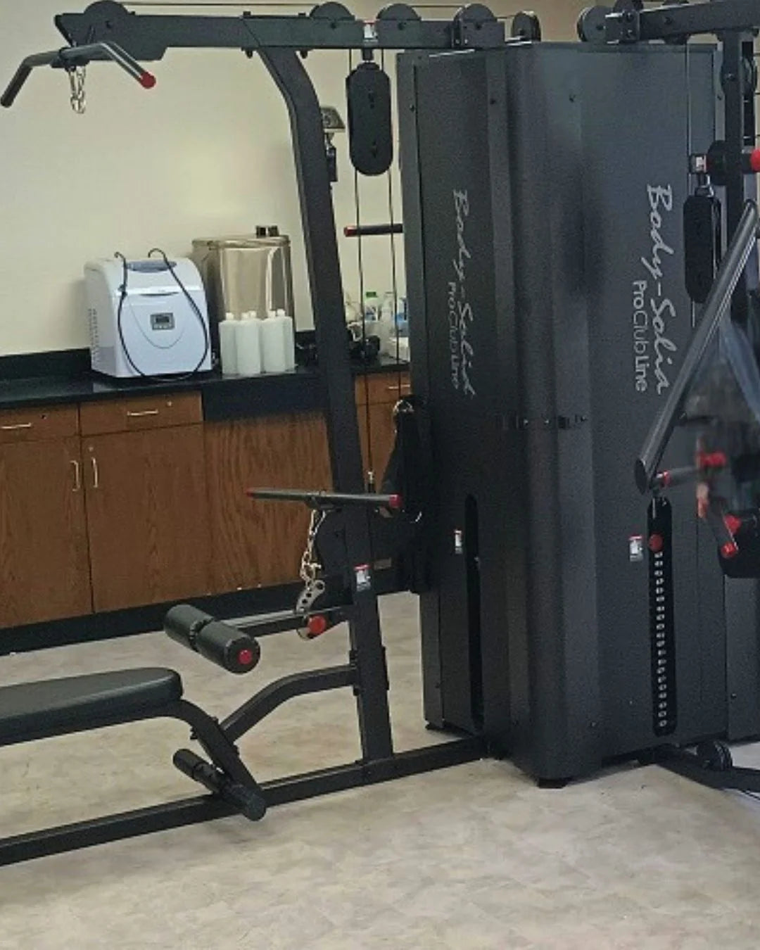 A gym room features a multi-station workout machine labeled Body Solid Pro Club Line. In the background, there are wooden cabinets, a white mini-fridge, a large silver container, and several bottles on the counter.
