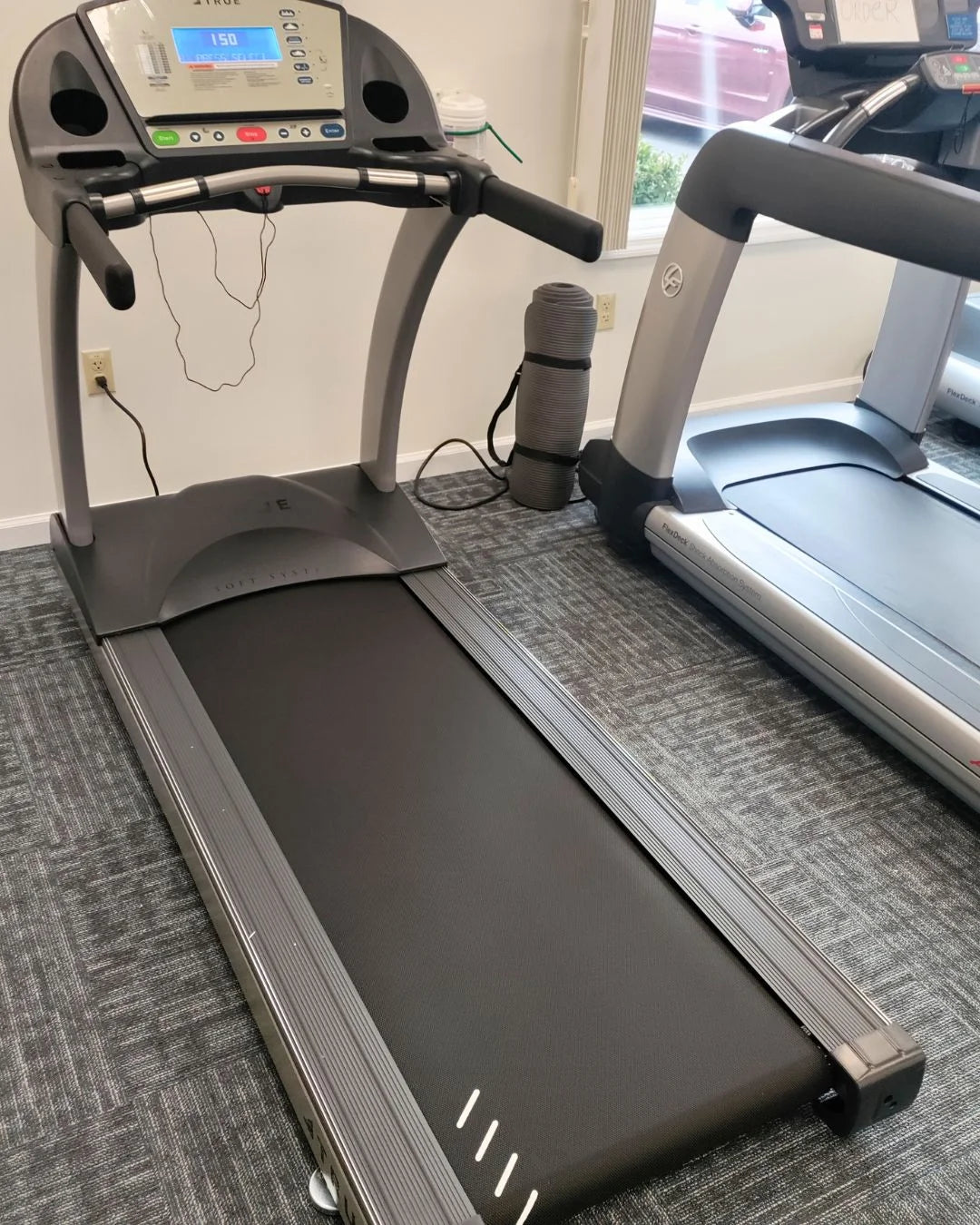 A treadmill in a gym room with a digital display showing a speed of 1.5 mph. Theres a rolled-up yoga mat and an adjacent treadmill nearby, all on a carpeted floor. Large windows are in the background.