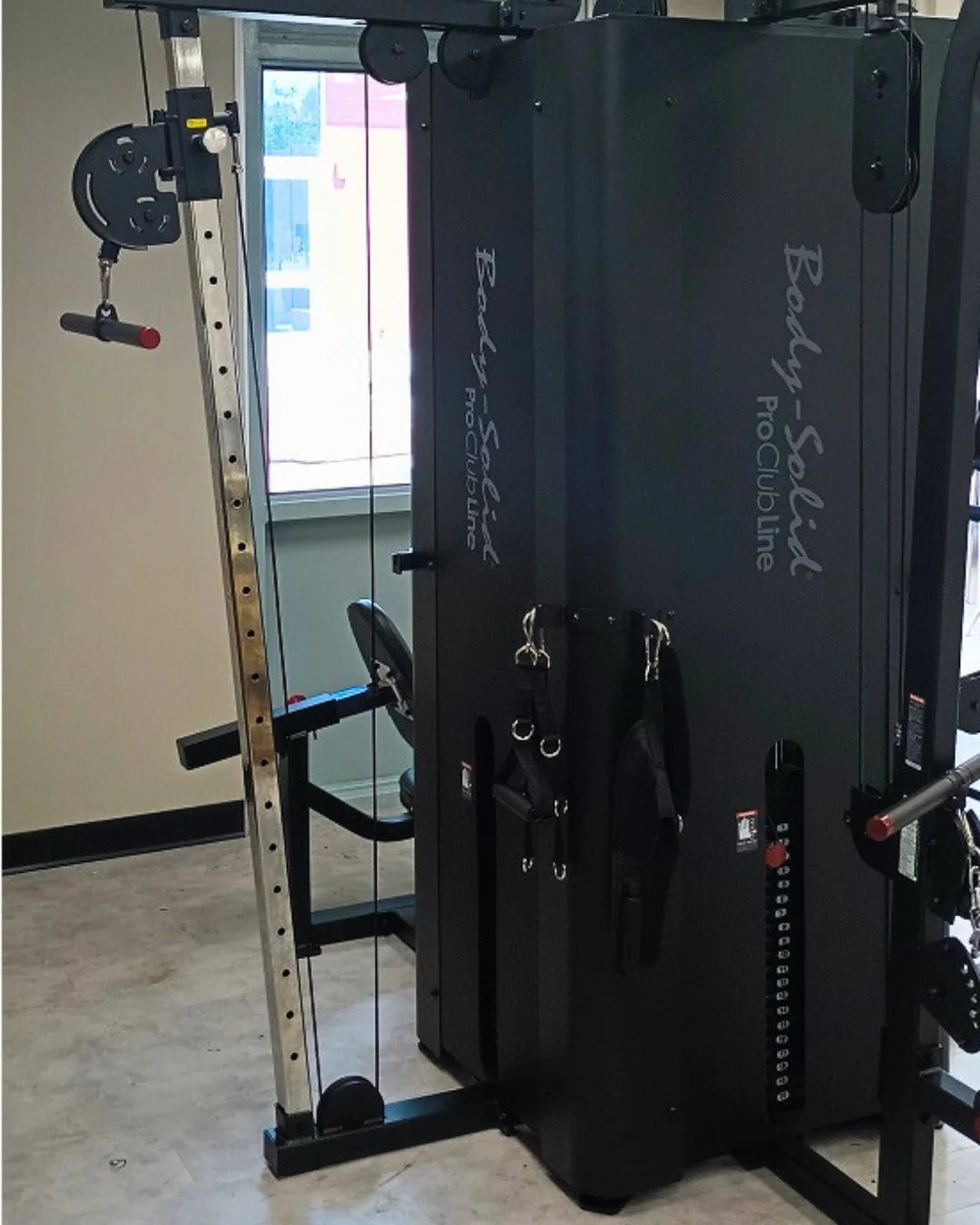 A gym features a multi-exercise machine with adjustable weights and cables. The equipment is black, with labeled Body Solid Pro Club Line. The room has a window, grey floor, and some wall-mounted attachments.