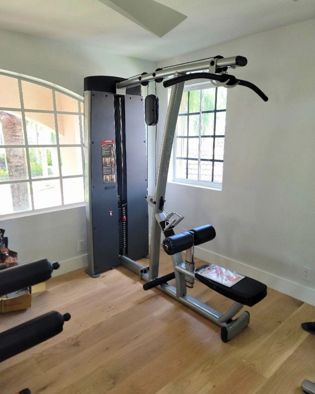 A home gym setup featuring a multifunctional weight training machine with pulleys and a leg extension attachment. The room has a large window, white walls, and wooden flooring, with another piece of gym equipment partially visible on the left.