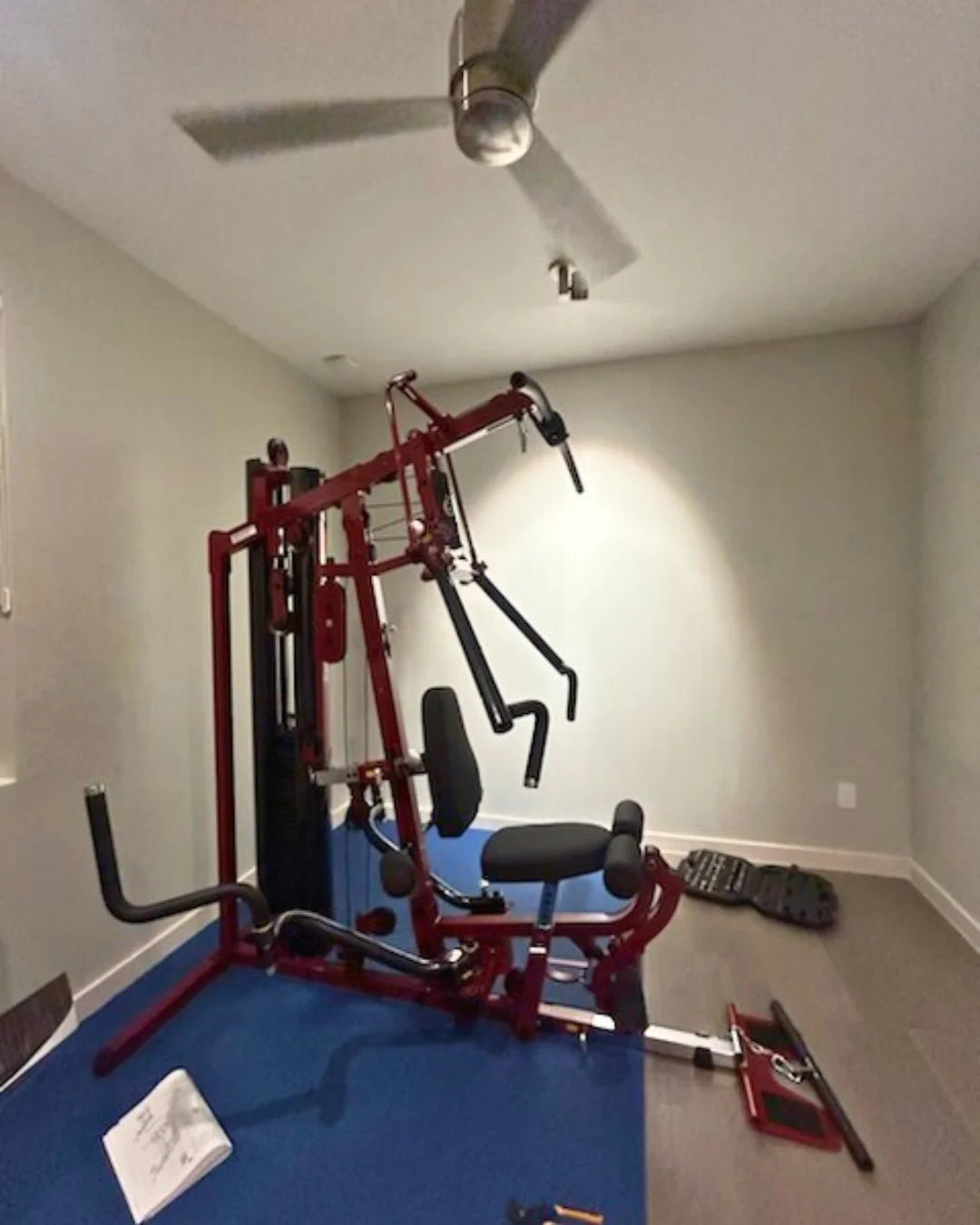 A home gym setup featuring a red multifunctional weight machine on a blue mat. The room has beige walls, a ceiling fan, and a small window to the left. A black mat and a few scattered items are visible on the floor.