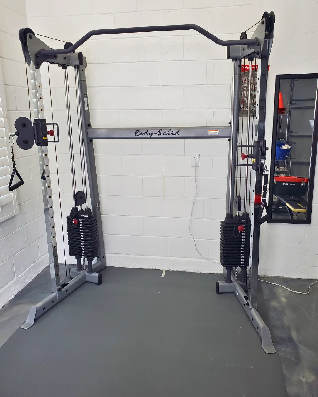 A cable crossover machine inside a gym with a white brick wall. It features two weight stacks, adjustable pulleys, and a pull-up bar. The floor is covered with gym mats, and theres a mirrored wall on the right with a display shelf.