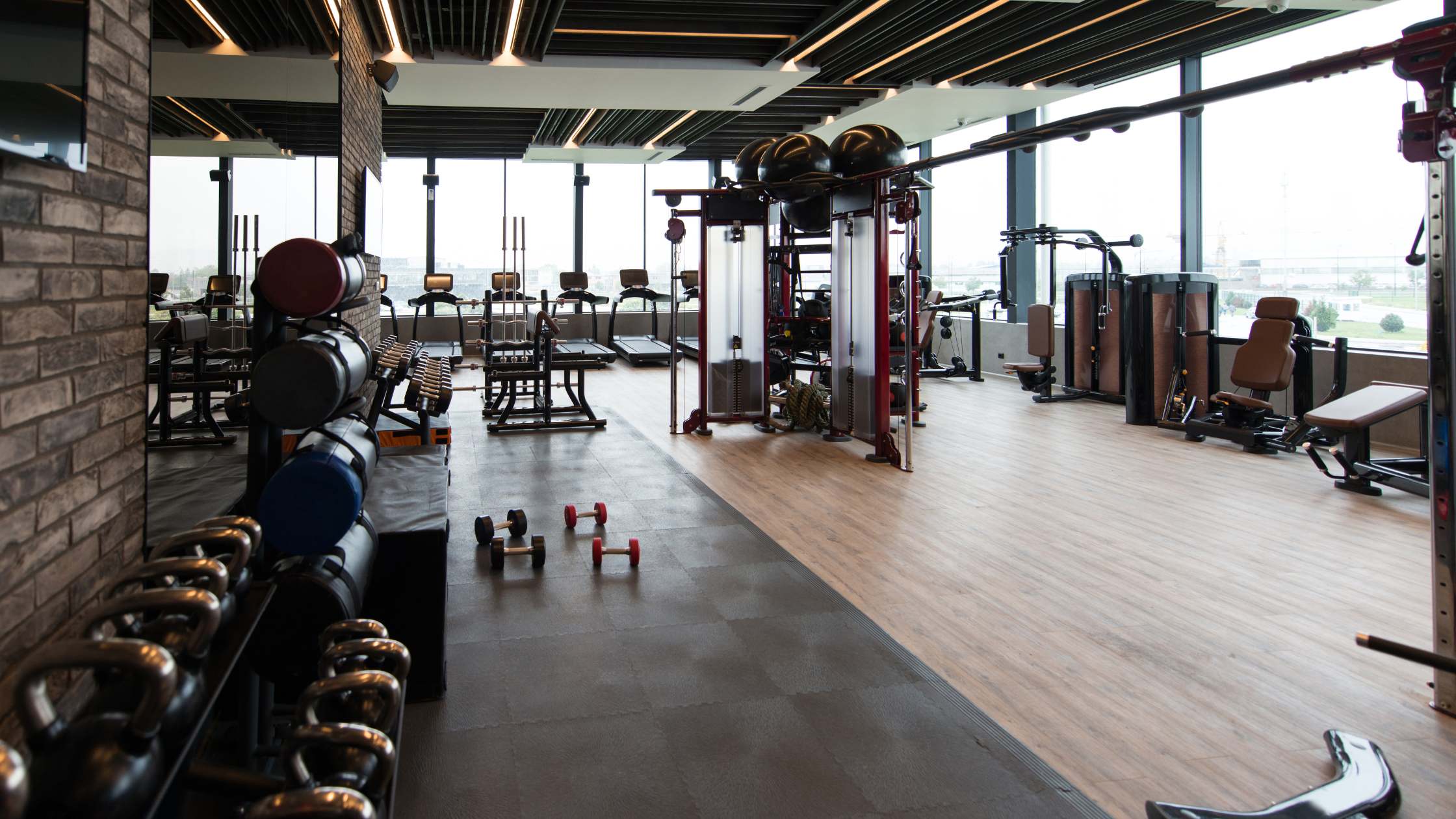 A brightly lit gym with a variety of workout equipment. There are free weights, kettlebells, benches, and exercise machines like elliptical trainers and cable machines. Large windows allow natural light to fill the space.