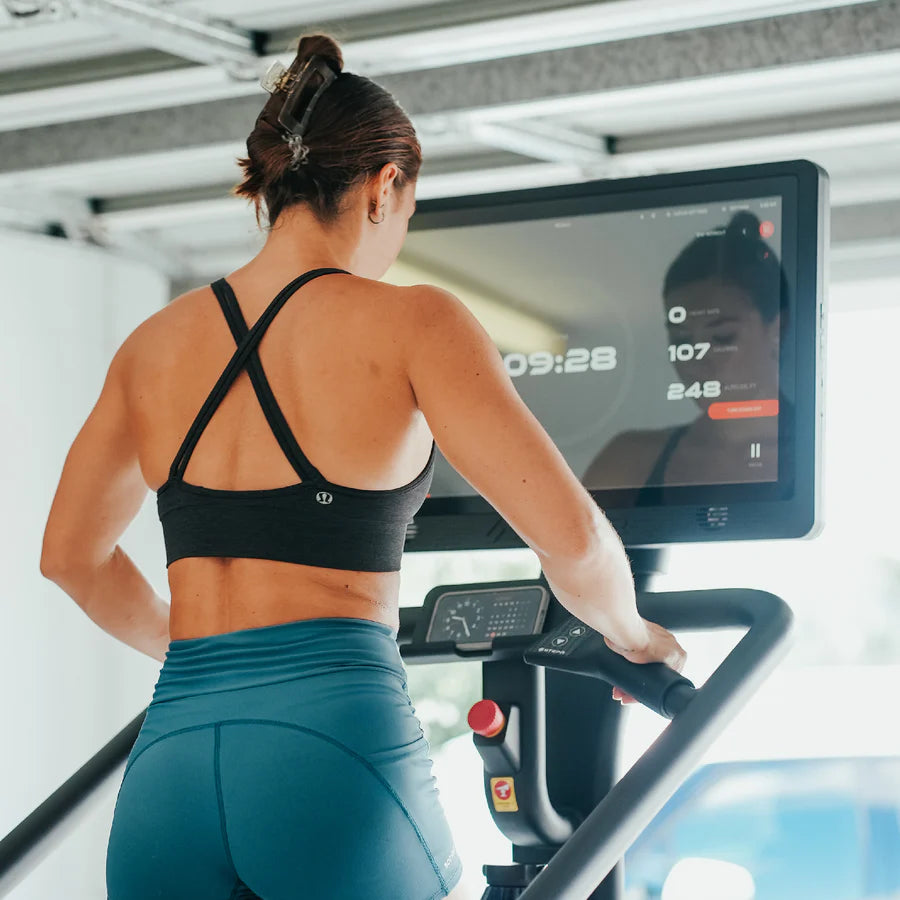 A woman in a black sports bra and teal leggings is using an exercise bike. She is looking at a digital screen displaying workout metrics like time, heart rate, and calories. The setting appears to be a bright indoor gym.