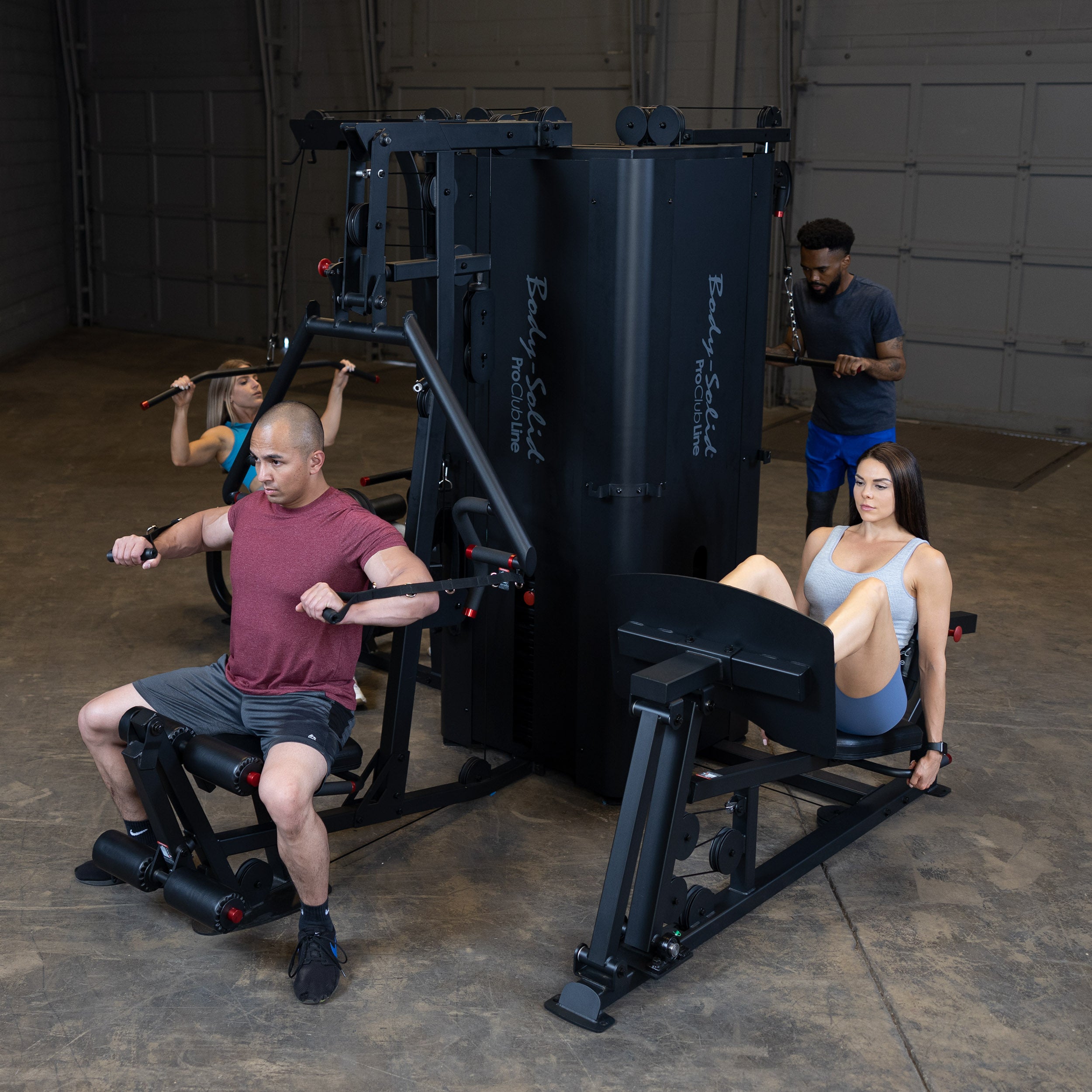Four people are exercising on a multi-station gym machine in a spacious room with a concrete floor. They are engaged in different workouts, including chest press, lat pull-downs, and leg presses.