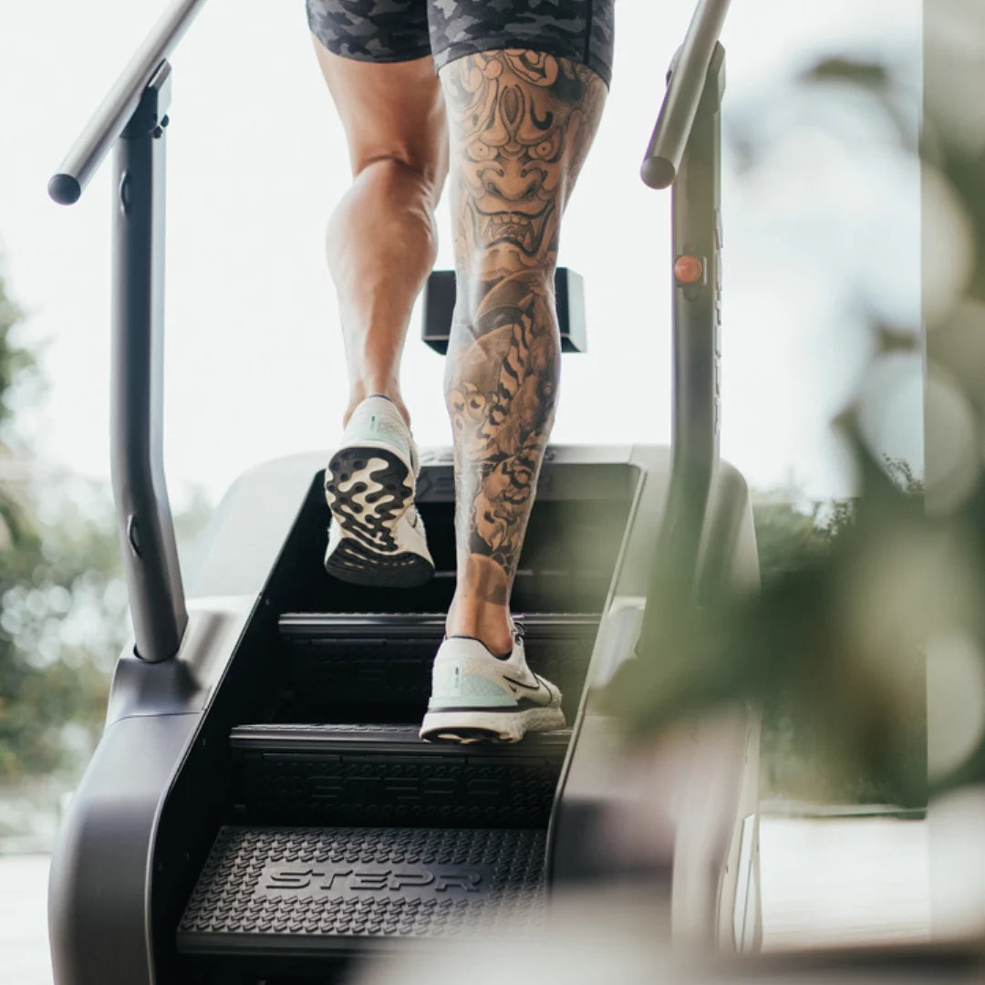 A person with a tattooed leg and wearing athletic shoes is using a stair-stepper machine. They are wearing dark camouflage shorts. The viewpoint is from behind, focusing on the tattoo and sneakers.