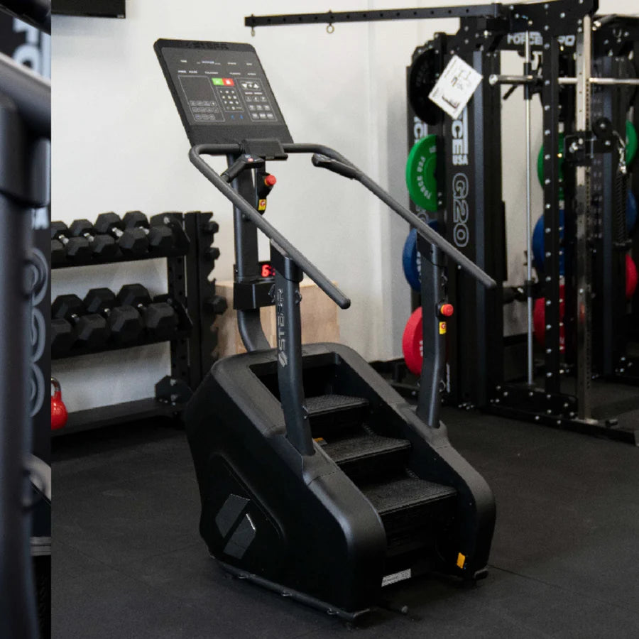 Stair stepper machine in a gym setting with a digital display panel. Its positioned near a rack of dumbbells and weightlifting equipment, including colorful weight plates. The floor is covered with black mats.
