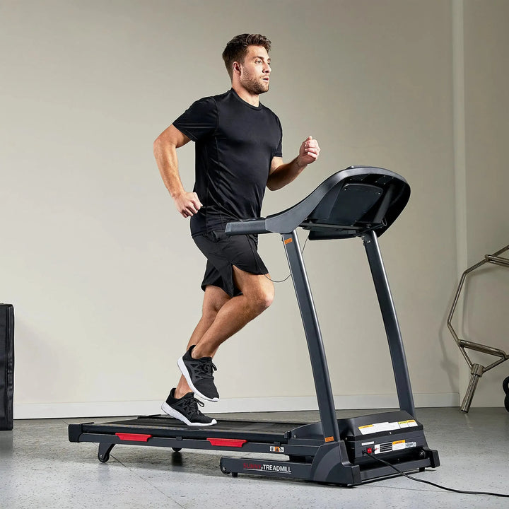 A man in a black T-shirt and shorts energetically jogs on a Sunny Health & Fitness Premium Folding Treadmill in a well-lit room. His black sneakers pound rhythmically as an exercise machine gleams under soft lights in the background.
