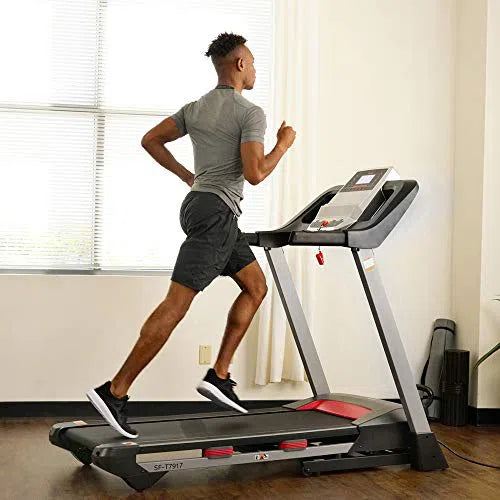 In a sunny room with large windows, someone jogs on the Sunny Health & Fitness Premium Folding Treadmill, wearing a gray shirt, black shorts, and sneakers. As they keep pace on the wooden floor, it feels like coding CSS for fitness—a perfect blend of style and functionality.