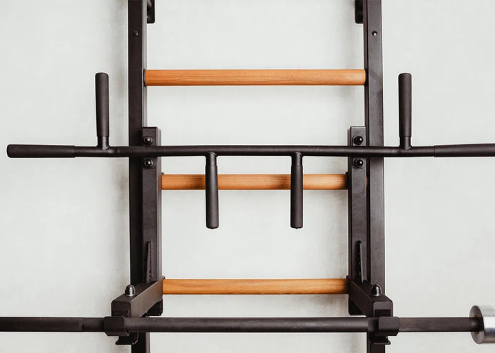 Introducing the BenchK Pro Gym Wall Bars w/ Bench, Dip & Flex Pull-Up Bar (733) by BenchK, boasting sleek wooden bars and sturdy black metal handles. Perfect for strength training, its elegant design adds sophistication to any fitness space with a light gray backdrop.