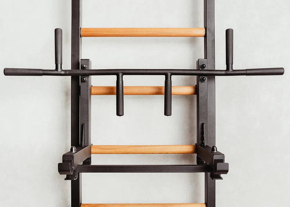 The BenchK Pro Gym Wall Bars (733) feature a sleek black metal frame and wooden rungs. This multifunctional unit includes a pull-up bar, various grips, a bench, and dip features, blending seamlessly into any home gym against a light-colored wall.