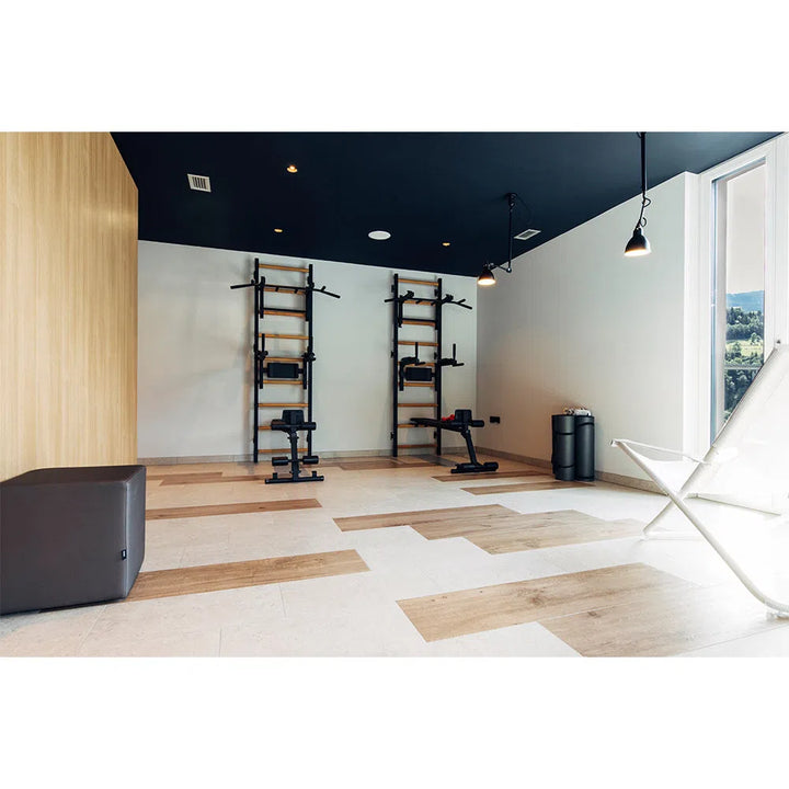 A modern home gym with light wood and white flooring includes BenchK Pro Gym Wall Bars w/ Bench, Dip & Flex Pull-Up Bar (733) on the back wall. A white lounge chair and a black cube seat complement the black ceiling and pendant lights.