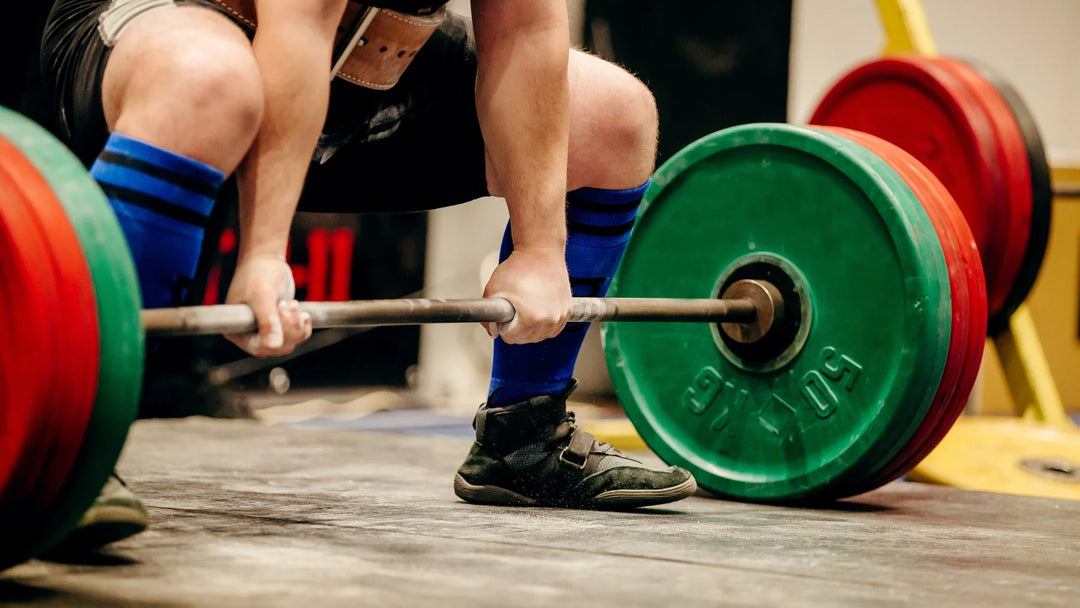 A person wearing a lifting belt and blue knee-high socks prepares to deadlift a barbell loaded with large red and green weight plates in a gym setting.