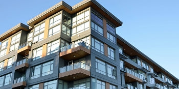 Modern apartment building with large glass windows and balconies, photographed from a low angle against a clear blue sky.
