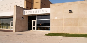 Exterior view of a modern beige building with large windows and a sign above the entrance that reads ATHLETICS. The sidewalk and a patch of grass are visible in front.