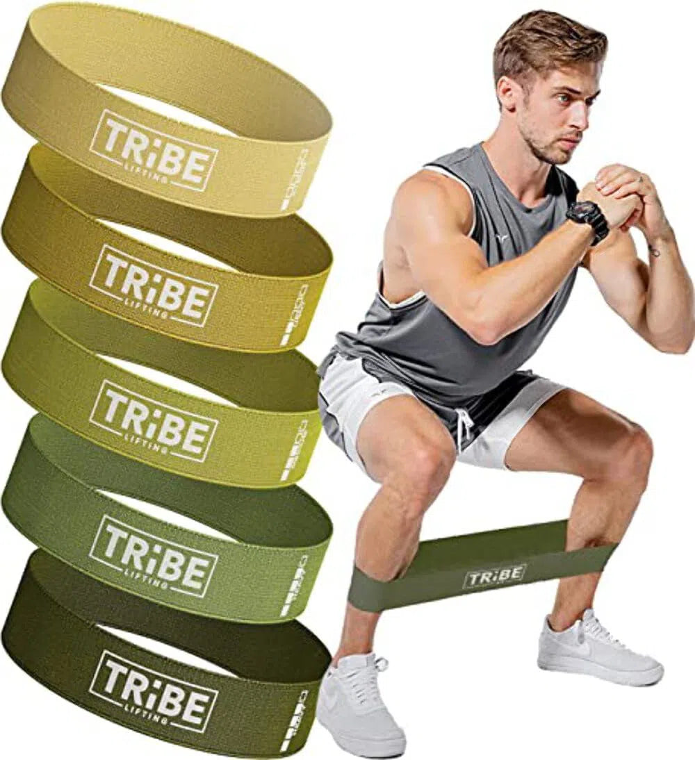 A man in athletic wear squats with a Tribe Fabric Resistance Band for Botty Workout around his thighs. Next to him, five yellow and green Tribe labeled bands are stacked. He wears a gray tank top and white shorts, highlighting his fitness commitment among neatly arranged workout gear.