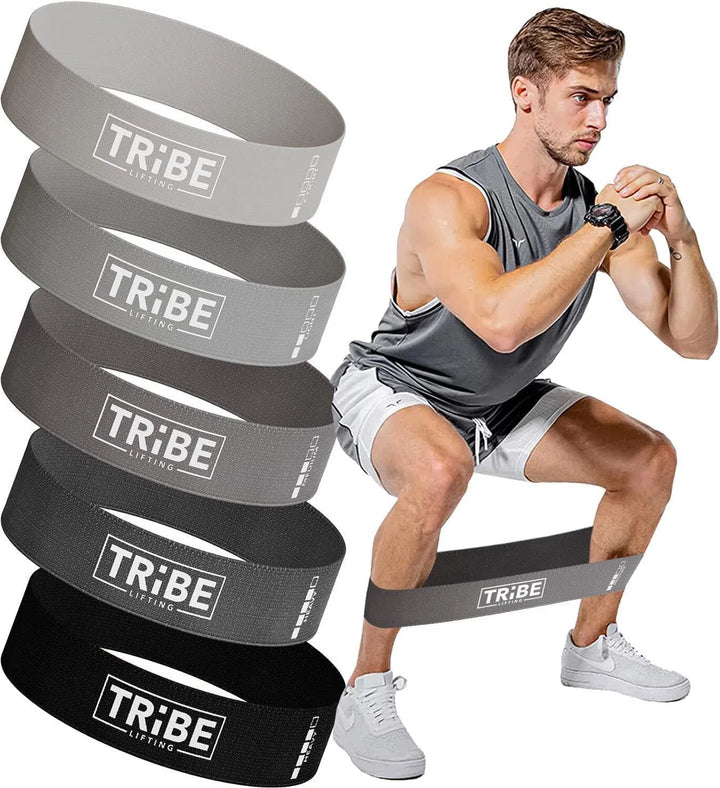 A man in a gray tank top and white shorts expertly demonstrates a squat using the Tribe Fabric Resistance Bands around his thighs. Beside him, five bands in shades from gray to black show varying resistance levels by Tribe, ideal for customizing workout intensity.