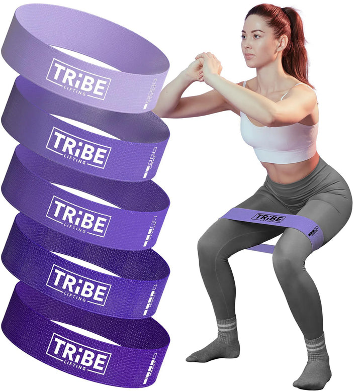 A woman expertly squats with a purple Tribe Fabric Resistance Band around her thighs. To her left, five colorful Tribe bands in purple and pink are on display. She wears a sleek white top and gray leggings, combining fitness with style effortlessly.