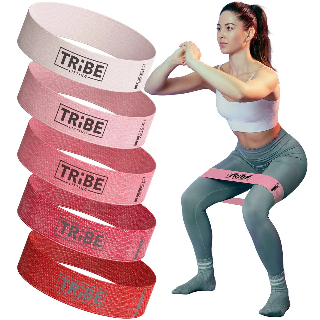 A woman in athletic wear squats with a pink Tribe Fabric Resistance Band around her thighs. Four more bands in shades of pink and red from the Tribe brand appear on the left, demonstrating their variety for great booty workouts without using typical product-related words.