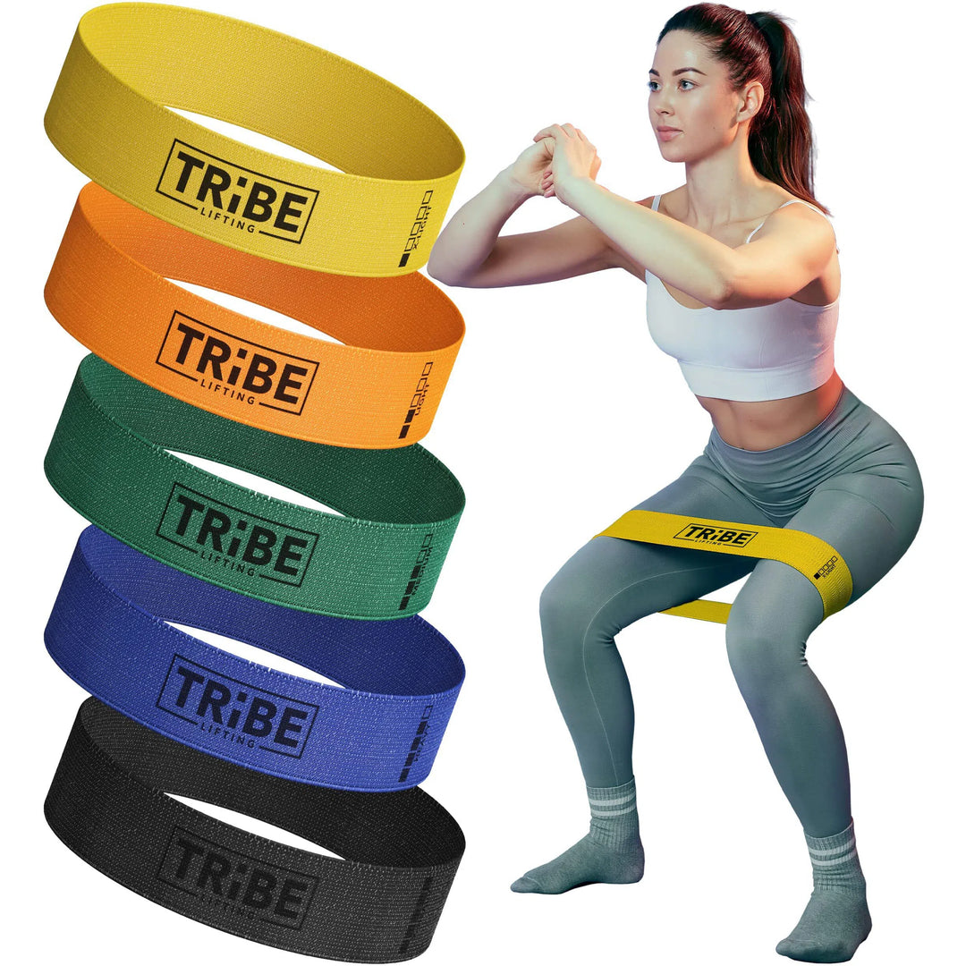 A woman in workout attire squats with a yellow Tribe Fabric Resistance Band around her thighs, surrounded by five more vibrant bands in orange, green, blue, black, and another yellow from the Tribe brand.