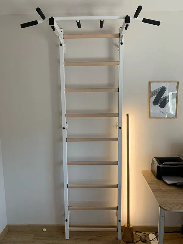 The BenchK Home Swedish Ladder w/ Pull-Up Bar (721), featuring a white wall-mounted design with wooden rungs, enhances the home gym near a wooden desk with a printer. Nearby, a tall floor lamp and framed abstract artwork complete the stylish setting.