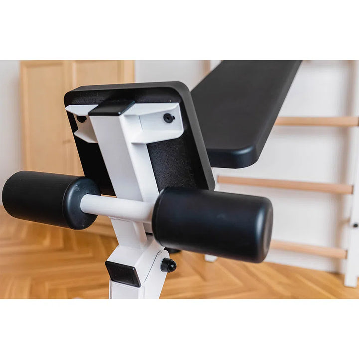 Close-up of the BenchK Pro Gym Swedish Ladder w/ Bench, Dip & Pull-Up Bar (723) in a gym. Focus is on the padded leg support and adjustable incline. The Swedish ladder complements the wooden gym bars on the polished floor.