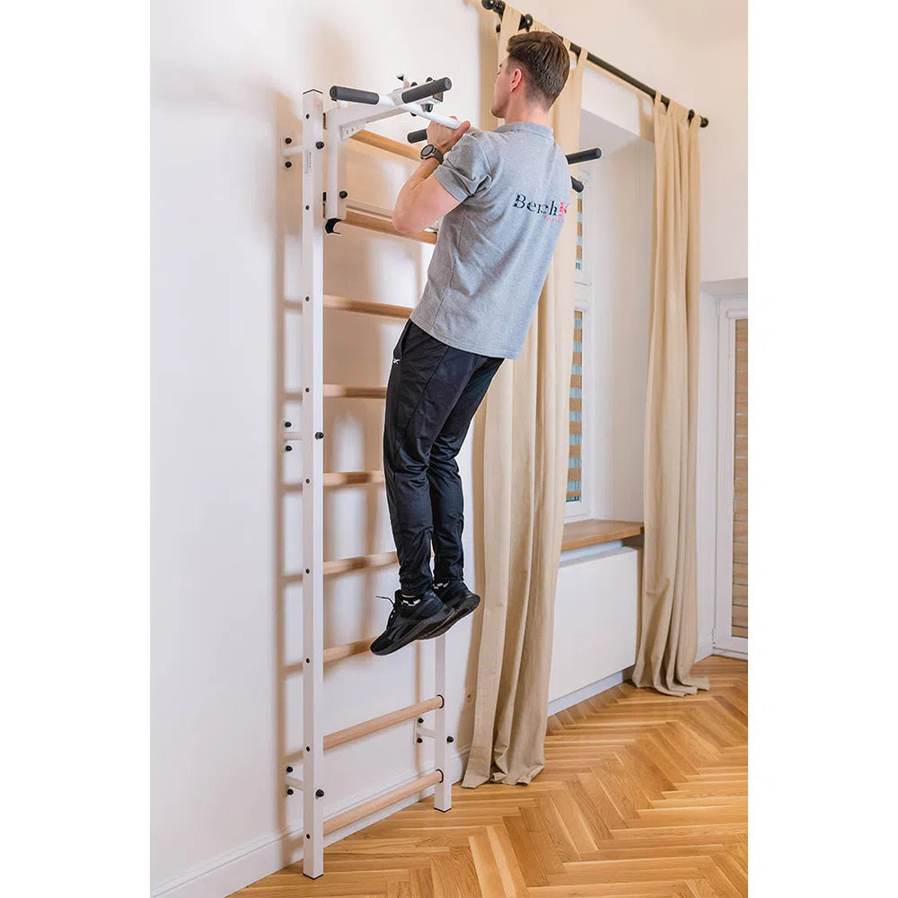 A person in a gray shirt and black pants performs a pull-up on BenchKs Home Wall Bars w/ Flex Pull-Up Bar (731) in a room with wooden flooring and beige curtains, ensuring their workout adheres to European safety standards.