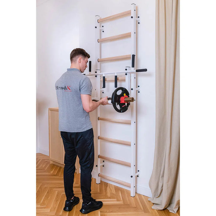 A man in a gray polo shirt and black pants stands by the BenchK Home Wall Bars with Flex Pull-Up Bar (731), placing a weight plate on it. The room, featuring a wooden floor and beige curtain, meets European safety standards.