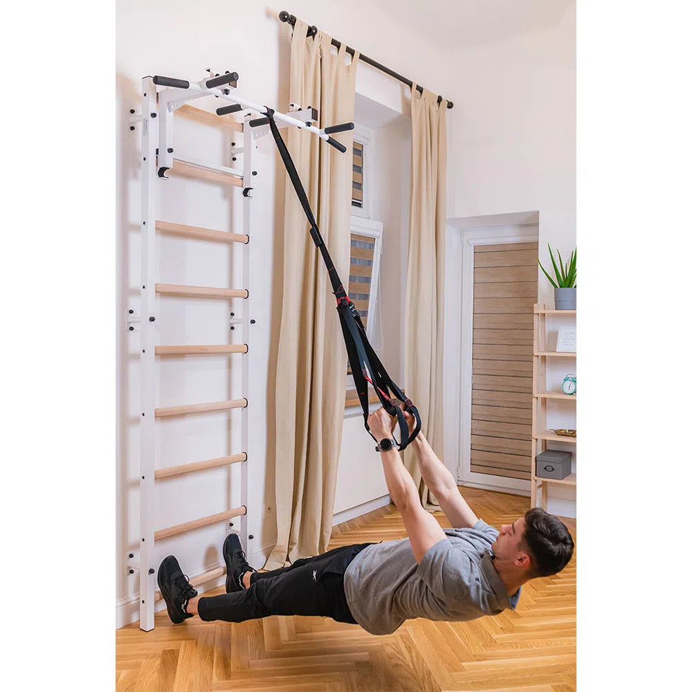 A person in gray and black workout gear uses a suspension trainer on the BenchK Home Wall Bars w/ Flex Pull-Up Bar (731), pulling bands in a plank position in a bright room with wooden floors, while ensuring European safety standards are met.