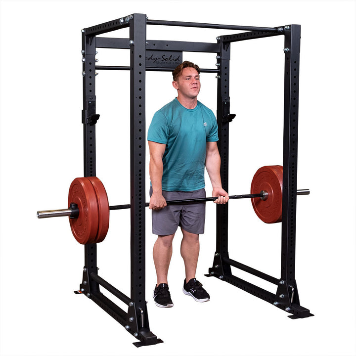 Man standing inside a Body-Solid GPR400 power rack holding a barbell loaded with red bumper plates at hip height for a rack pull