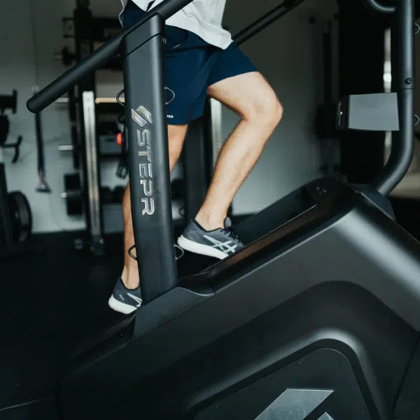 A person in navy shorts and sneakers briskly exercises on a black STEPR+ Stair Climber, highlighting its space-saving design ideal for home fitness. While the upper body remains unseen, gym equipment and weights create a vibrant backdrop.