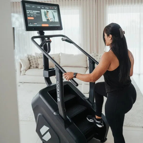 In a bright living room, a woman in black workout attire with a smartwatch uses the space-saving STEPR+ Stair Climber by STEPR. She follows a fitness program on its screen, with a couch and sheer curtains in the background.