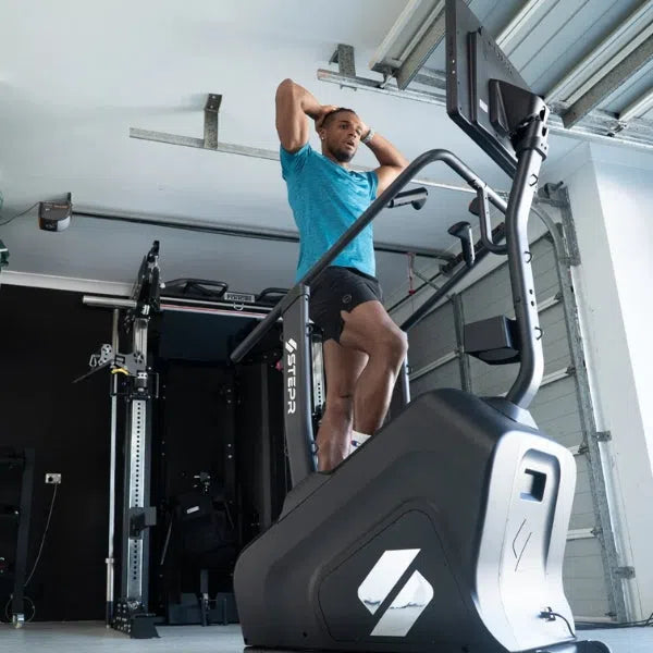 In his home garage gym, a man in a blue shirt and black shorts exercises on a STEPR+ Stair Climber by STEPR. With hands on his head, he’s surrounded by fitness equipment in this modern, industrial-style space that boasts a sleek, space-saving design.