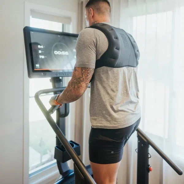 A tattooed individual in a gray t-shirt and black shorts uses the STEPR+ Stair Climber, checking workout stats on its digital screen and enjoying natural light. The STEPR+ design is compact, making it ideal for any home gym setup.