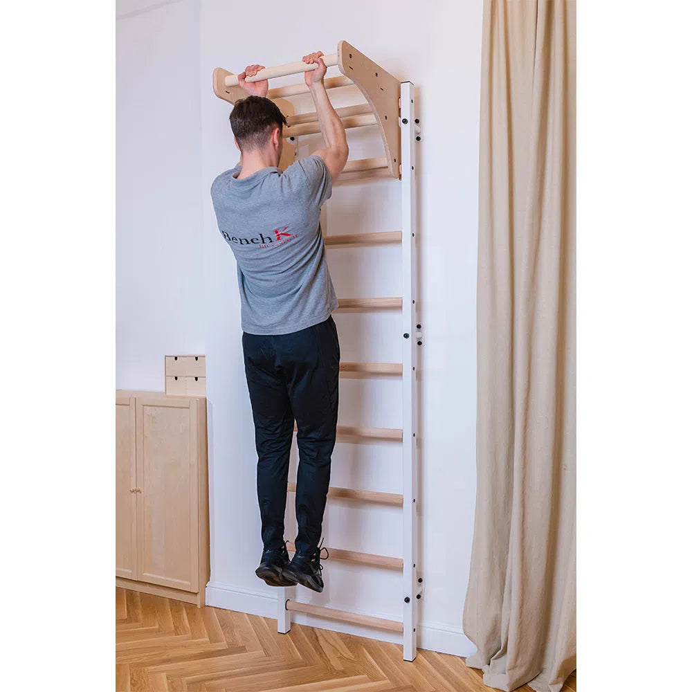 A person in a gray T-shirt and black pants is using the BenchK Home Stall Bars with Wooden Pull-Up Bar (711), part of their home gym, mounted on a wall in a room with wooden floors and a beige curtain.