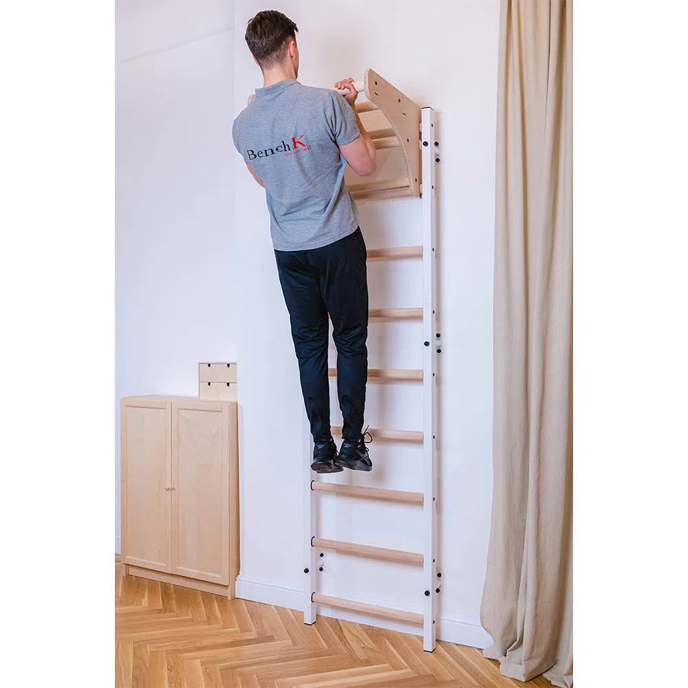 A person wearing a gray T-shirt and black pants stands on the BenchK Home Stall Bars w/ Wooden Pull-Up Bar (711) in their versatile home gym. The room features wood flooring, a light-colored cabinet, and curtain in the background.