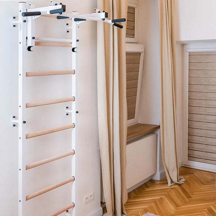 The BenchK Home Wall Bars w/ Flex Pull-Up Bar (731) feature a sleek white frame and wooden climbing ladder, set in a minimalist corner with beige curtains, herringbone wood flooring, and a radiator beneath the window. This setup meets European safety standards.