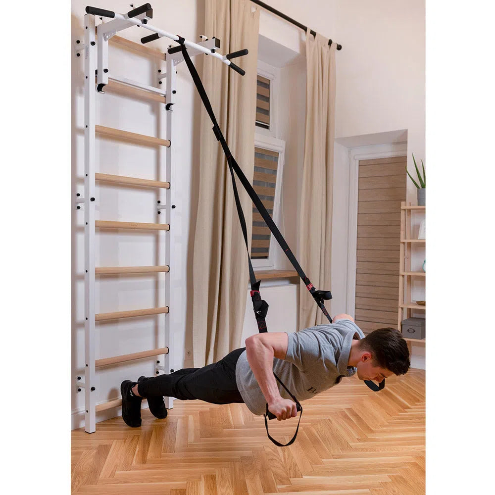 A person does push-ups with suspension training straps on the BenchK Home Wall Bars w/ Flex Pull-Up Bar (731). The European safety-compliant room features wooden flooring, light-colored curtains, and a potted plant in the corner.