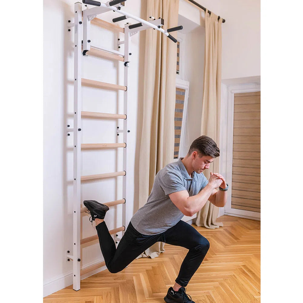 A man in a gray t-shirt and black pants does lunges with the Flex Pull-Up Bar on BenchKs Home Wall Bars (731). The room, adhering to European safety standards, includes wooden floors, light-colored walls, and curtains beside a window.