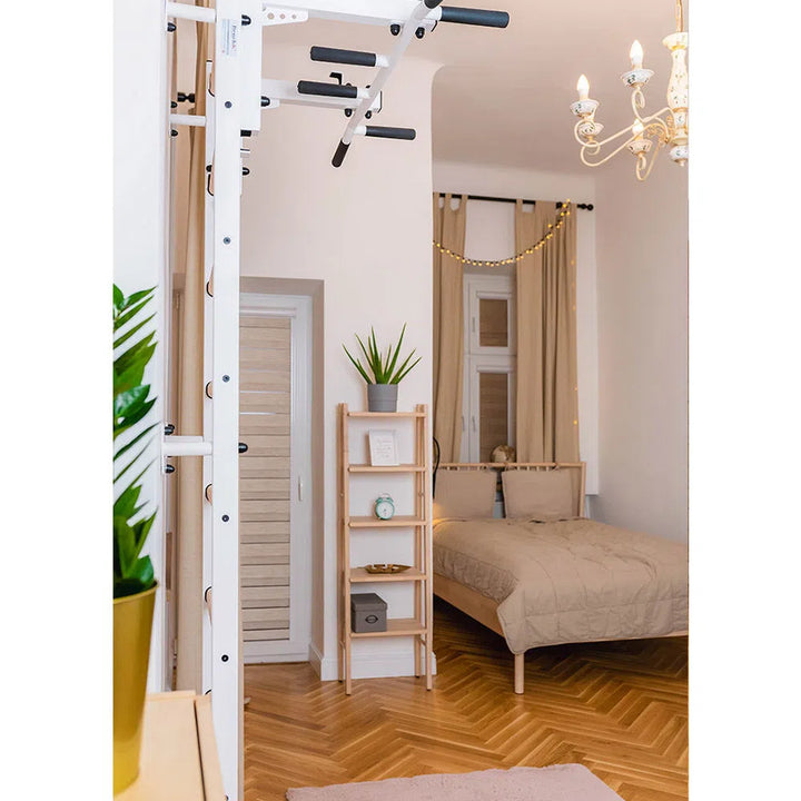 A cozy bedroom includes a bed with beige bedding, a small wooden shelf with plants and books, and BenchKs 731 Home Wall Bars w/ Flex Pull-Up Bar that meets European safety standards. A chandelier elegantly hangs from the ceiling, while a door is visible in the background.