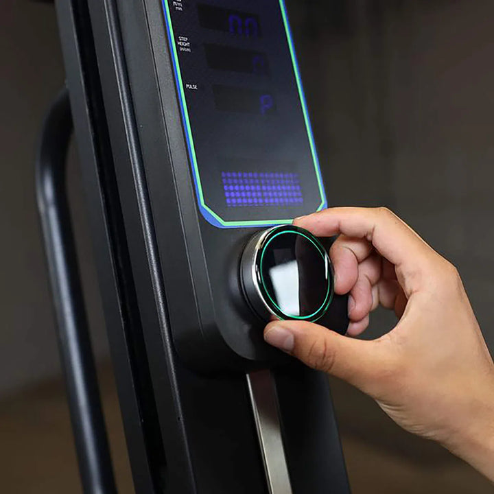 A user tweaks a digital knob on the Body-Solid Endurance Mountain Climber Machine (CL300), featuring illuminated blue indicators. The background is blurred, showcasing interaction with the electronic device crafted for customizable resistance and a comprehensive full-body workout.