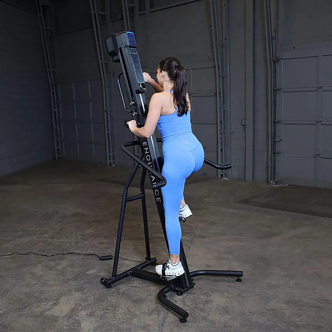 In a spacious room with gray walls and concrete flooring, a person in a blue workout outfit uses the Body-Solid Endurance Mountain Climber Machine (CL300), facing away as they climb with one foot on a pedal and both hands on handles, enjoying its adjustable resistance for an optimized full-body workout.