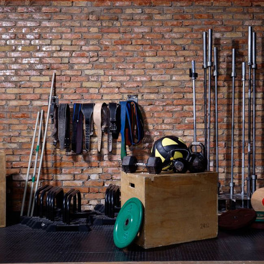 A gym with various equipment against a brick wall. There are barbells, weightlifting belts, dumbbells, a medicine ball, kettlebells, and a plyometric box. Weight plates and resistance bands are also visible on the floor and rack.