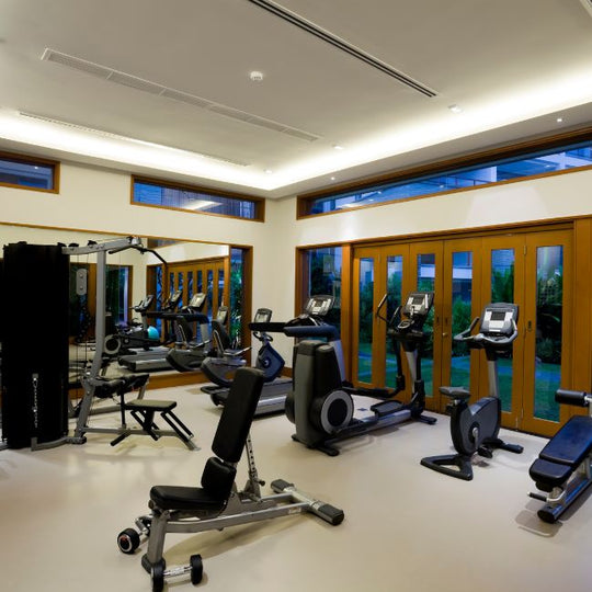 A modern gym with large windows features various exercise equipment, including treadmills, stationary bikes, and weight machines. Light-colored walls and flooring create an open and inviting atmosphere.