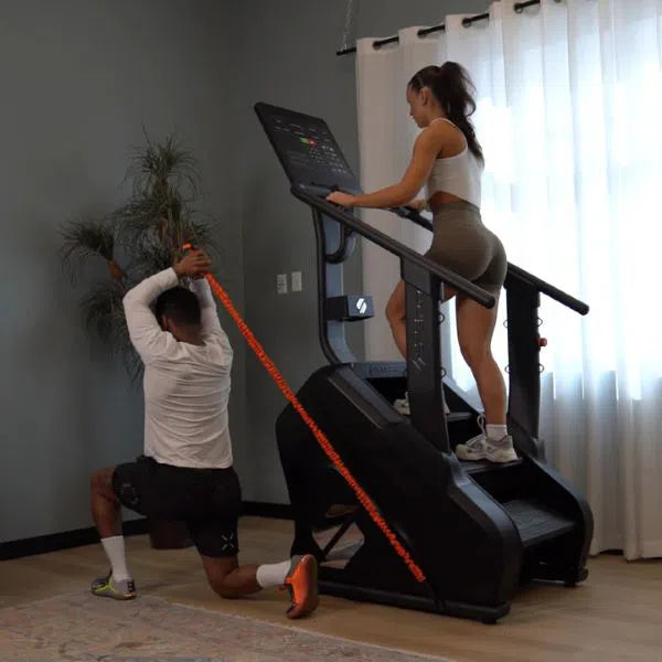 A woman in workout gear uses the STEPR PRO Classic Stair Climber indoors, while a man in a white shirt and shorts kneels, performing high-intensity training with an overhead elastic band. The room has gray walls, a plant, and sheer white curtains.