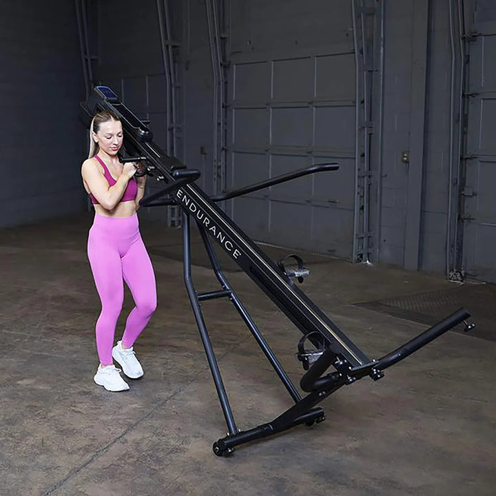 A person in a pink workout outfit adjusts the adjustable resistance on a Body-Solid Endurance Mountain Climber Machine (CL300) in a spacious warehouse, optimizing their full-body workout.