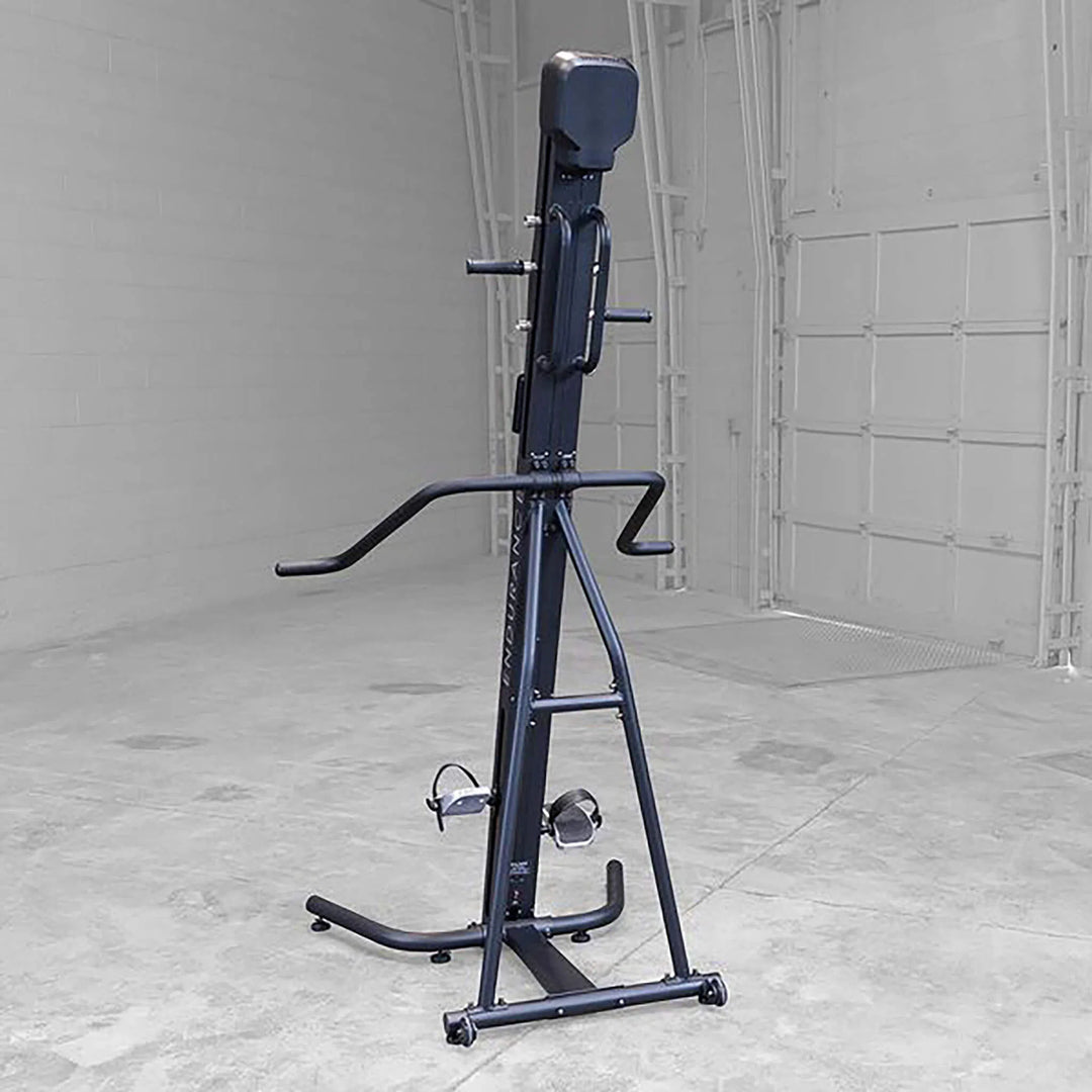 The Body-Solid Endurance Mountain Climber Machine (CL300) is set against an industrial backdrop with a white garage door and concrete floor. It features adjustable resistance, handlebars, foot pedals, and a cushioned seat back for a full-body workout.