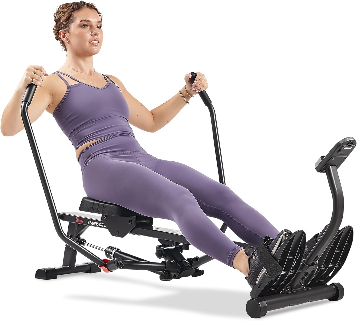A woman in a purple workout outfit uses a rowing machine, exercising indoors with her hands gripping the handles and feet secured on the footrests.